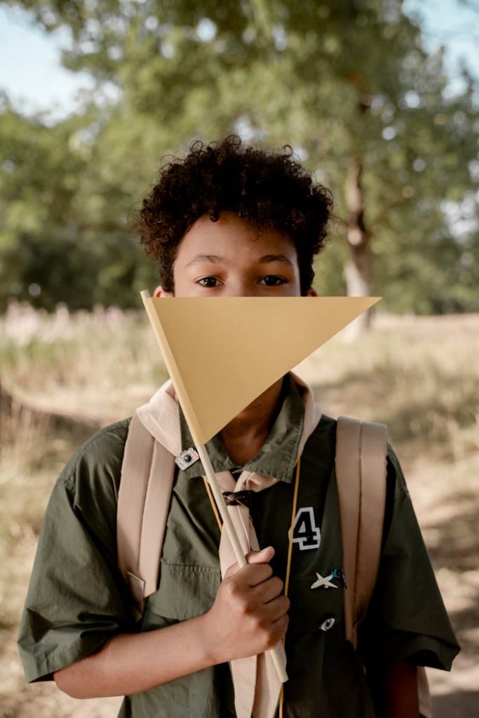 A young boy scout with afro hair holds a triangular flag in a sunny, outdoor setting.