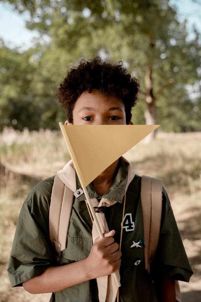 A young boy scout with afro hair holds a triangular flag in a sunny, outdoor setting.